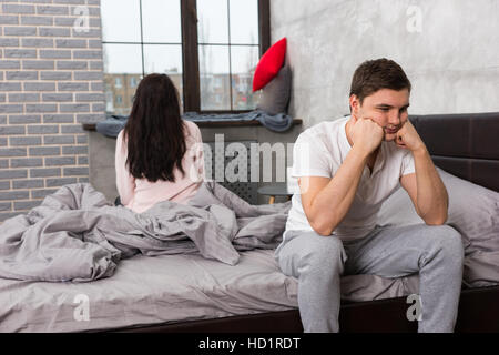 Young man is leaning on his hands while sitting sadly on bed, woman is sitting on bed and looking out the window in the background Stock Photo