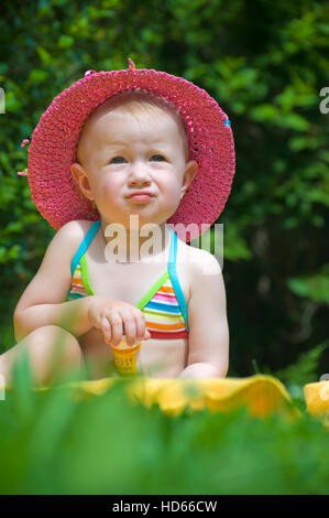Infant girl wearing a sunhat and a bikini sitting on a meadow and playing with soap bubbles Stock Photo