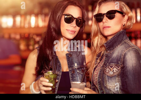 Two gorgeous women with refreshing drinks having fun in the outdoor bar, wearing stylish sunglasses and enjoying beautiful sunset Stock Photo