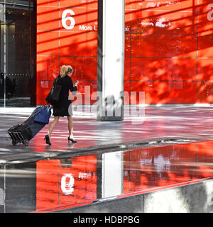 Smart woman walking with luggage trolley in pedestrianised street in More London holding mobile phone red water reflection in water feature Stock Photo