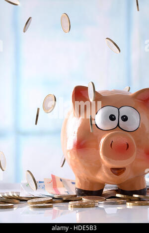 Concept savings with coins falling on piggy bank with savings billboard and stacked coins on reflective white glass table and blue windows background.