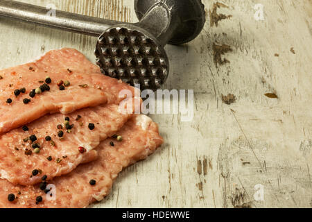 Three raw, broken chops sprinkled with peppercorns and salt crystals, meat tenderizer on an old wooden kitchen table. Close, horizontal view. Stock Photo