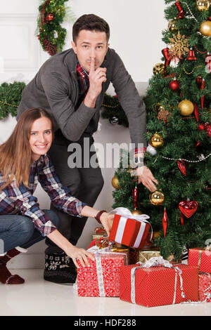Happy parents with finger on lips put christmas gifts for children under decorated tree Stock Photo
