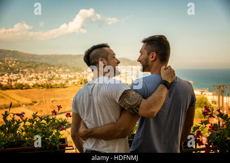 Back view of homosexual couple embracing nd looking at each other on background of resort. Stock Photo