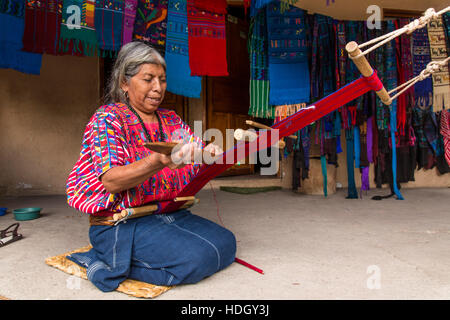 An older, grey-haired Mayan woman weaves fabric on a backstrap loom while kneeling on the floor of her home in Santa Catarina Palopo, Guatemala. Stock Photo