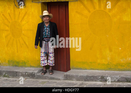 An older Mayan man in traditional dress of San Pedro la Laguna, Guatemala, stands in front of brightly painted house with a red door and yellow sun de Stock Photo