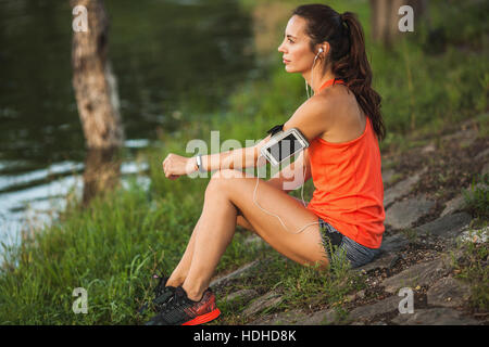 Side view of young woman sitting on rock by pond while listening to music at park Stock Photo