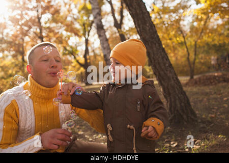 Father blowing bubbles with baby boy in park during autumn Stock Photo