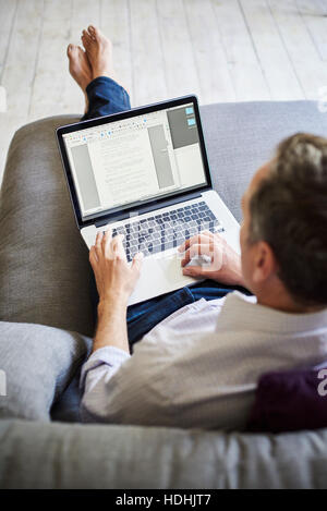 A man seated on a sofa, using a laptop computer, over the shoulder view. Stock Photo