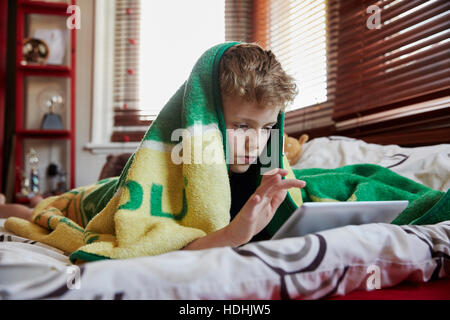 A boy with a towel over his head lying on his bed, using a digital tablet. Stock Photo