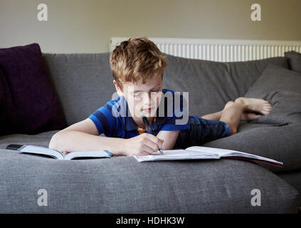 A family home. A boy lying on his front on a sofa doing his homework, writing in an exercise book. Stock Photo