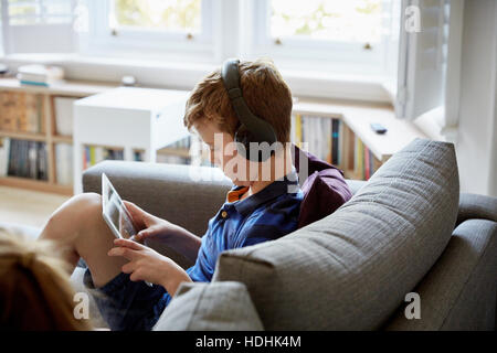 A family home. A boy on a sofa, using a digital tablet, wearing headphones. Stock Photo