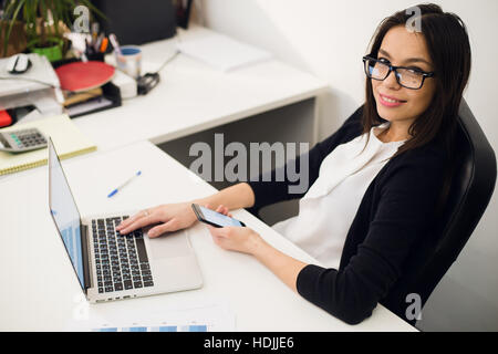 Good business talk. Cheerful young beautiful woman in glasses talking on mobile phone and using laptop with smile while sitting at her working place Stock Photo