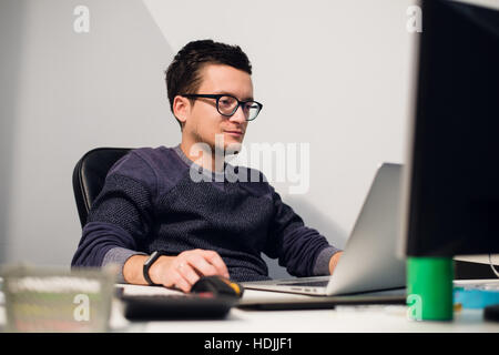 Busy working. Pensive young handsome man wearing glasses on laptop while sitting at his work place Stock Photo
