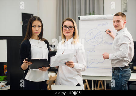 Two beautiful office workers having conversation while man colleague drawing business strategy on flip chart. Stock Photo