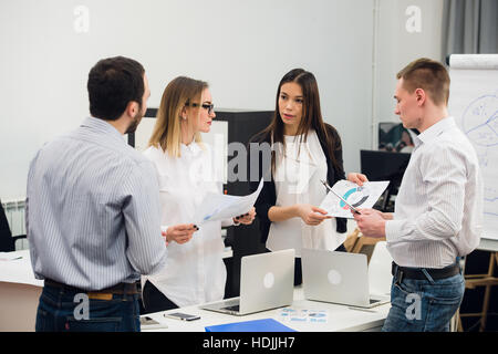 Four young business people working as a team gathered around laptop computer in an open plan modern office Stock Photo