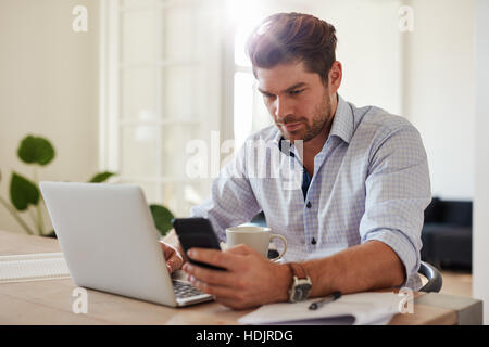 Shot of young man working with laptop and mobile phone at his home office. Handsome male sitting at table holding smartphone and using laptop computer Stock Photo