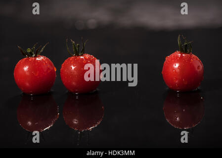 Three tomatoes with water drops lie on dark background, reflected on black background Stock Photo