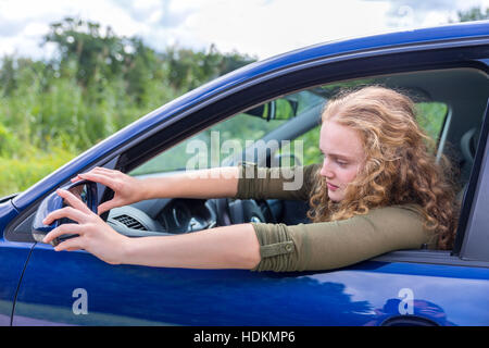 European woman adjusting side mirror of blue car in nature Stock Photo