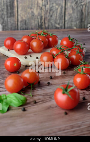 Scattered cherry tomatoes with pepper and leaves of basil on the wooden, brown table with the light wooden board. Stock Photo