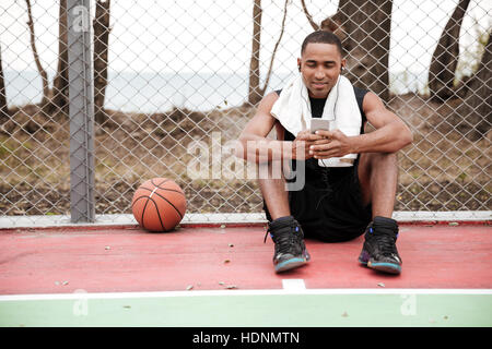 Picture of young happy african basketball player sitting in the park with towel near basketball and chatting while listen to music. Look at phone. Stock Photo