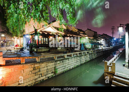 Retail services and street food cafes along shopping street and canal in ancient Suzhou city downtown at sunset. Bright street lamps illuminate histor Stock Photo