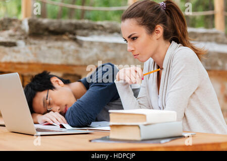 Concentrated young woman sitting and studying near man sleeping on the desk Stock Photo