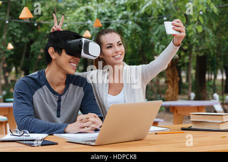 Cheerful young couple using virtual reality glasses and making selfie with smartphone outdoors Stock Photo