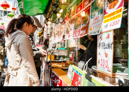 Japan, Kobe, Nankinmachi, Chinatown. Side view of young Japanese couple, buying from counter of Chinese takeaway with many photo menu cards hanging. Stock Photo