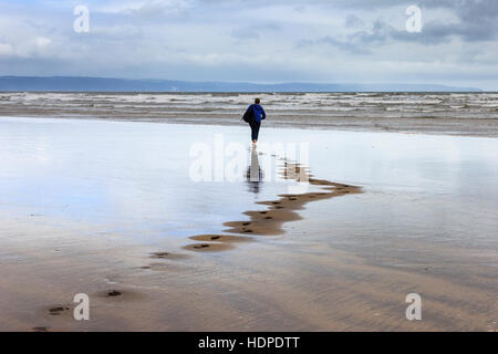 One woman walking barefoot towards the sea on a windswept beach leaving footprints in the sand, Westward Ho!, Devon, England, UK Stock Photo