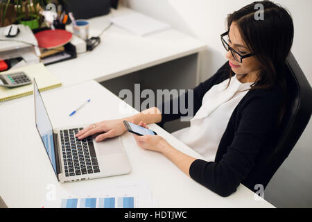 Good business talk. Cheerful young beautiful woman in glasses talking on mobile phone and using laptop with smile while sitting at her working place Stock Photo