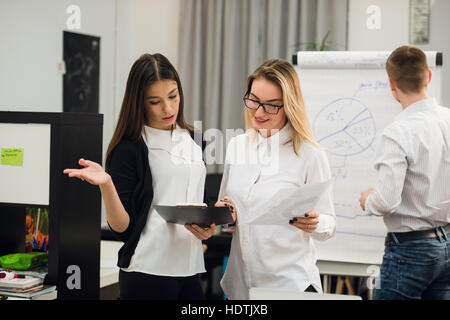 Two beautiful office workers having conversation while man colleague drawing business strategy on flip chart. Stock Photo