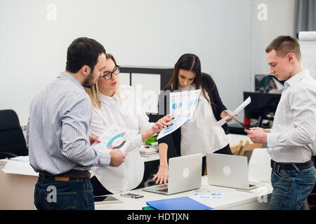 Four young business people working as a team gathered around laptop computer in an open plan modern office Stock Photo