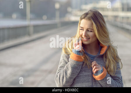 Happy woman with a lovely smile standing in her coat on a sea promenade in winter looking at the camera with a joyful expression and her hair blowing  Stock Photo