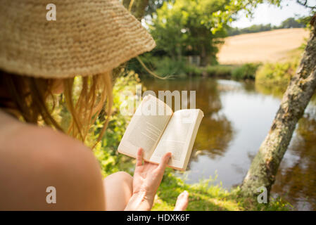 Woman wearing a hat reading a book on holiday in English countryside by a river Devon England Stock Photo