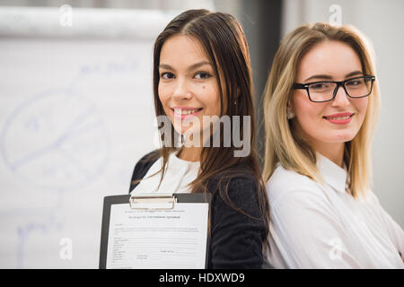 Two businesswomen coworkers standing in an office and smiling positively at the camera while holding folder of paperwork Stock Photo