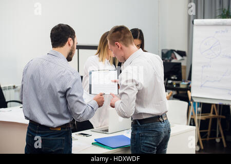 Four young business people working as a team gathered around laptop computer in an open plan modern office Stock Photo