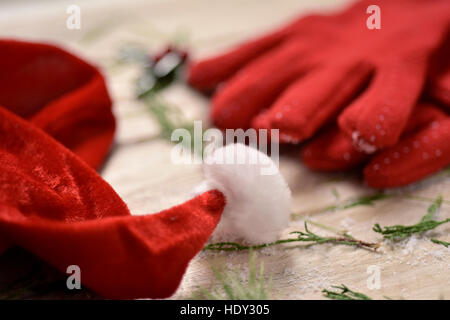 closeup of a santa hat, a pair of cozy red gloves, some twigs of pine tree and a twig of holly in the background, on a rustic wooden table Stock Photo