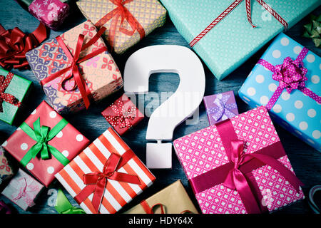 high-angle shot of many gifts wrapped in nice papers and tied with ribbons of different colors and a white three-dimensional question tag in the cente Stock Photo
