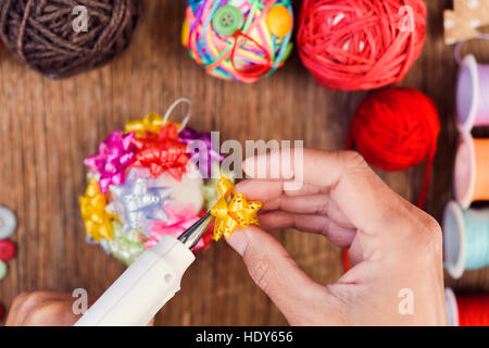 high-angle shot of a young man making a handmade christmas ball with ribbon bows of different colors, using a hot glue gun, and a pile of different ba Stock Photo