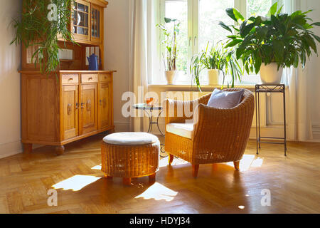 Sunny living room in warm colors with cozy wicker armchair and stool, vintage cupboard, plants and large windows, nostalgic Stock Photo