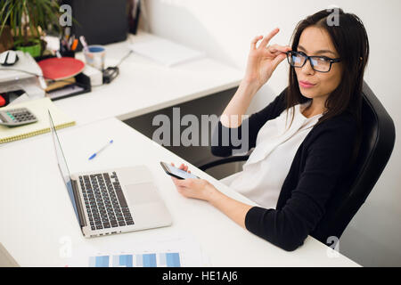 Good business talk. Cheerful young beautiful woman in glasses talking on mobile phone and using laptop with smile while sitting at her working place Stock Photo
