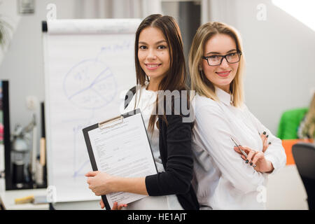 Two businesswomen coworkers standing in an office and smiling positively at the camera while holding folder of paperwork Stock Photo