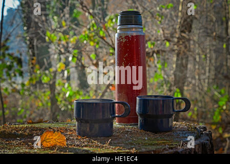 Red worn out thermos bottle with two blue cups on a tree trunk in autumn Stock Photo