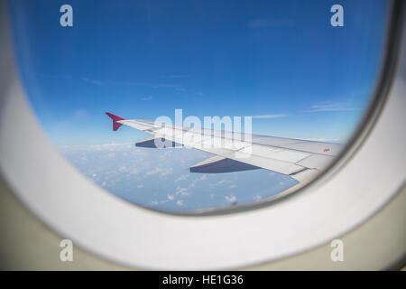 View through a window of an left airplane wing aircraft flying above the clouds in a blue sky. Stock Photo