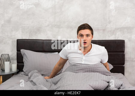 Frightened man woke up from a nightmare sitting in stylish bed with grey colors and near bedside table with candles in a bedroom in loft style Stock Photo