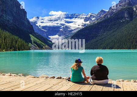 Two tourists relax on the boardwalk beside Lake Louise with a view of Mount Victoria in the distance Banff National Park Alberta Stock Photo