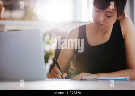 Young woman working at her desk taking notes. Asian businesswoman with laptop writes on a document at her office. Stock Photo