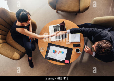 Top view of young business partners shaking hands on deal at office. Documents and laptop on table showing statistics and graphics. Stock Photo