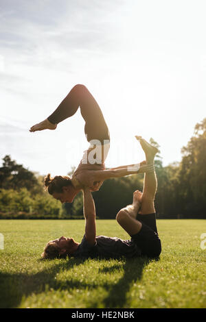 Vertical shot of healthy young couple doing acrobatic yoga on grass. Man and woman exercising in pair at the park. Stock Photo